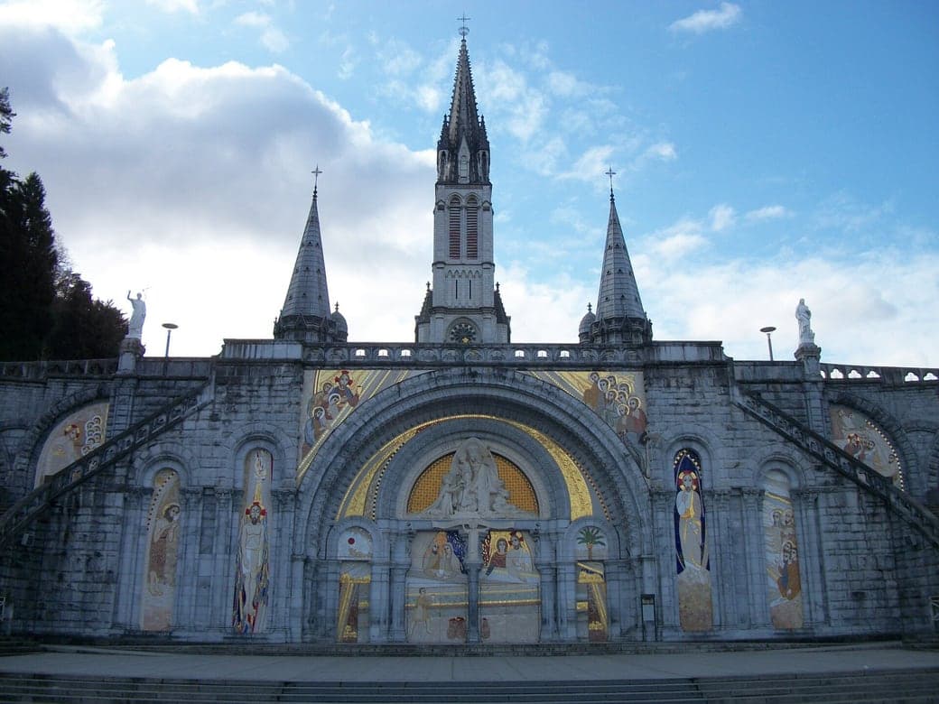 La façade de la basilique du Rosaire à Lourdes, mosaïques et flèches sous un ciel clair.
